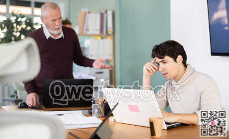 Senior man head of department shouts at guy subordinate who is working at laptop