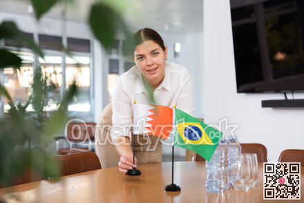 Positive young woman putting flag of Bahrain on the table with flag of Brazil in conference room