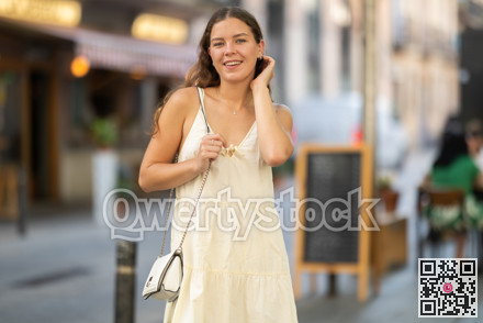 Cheerful girl in white dress walking along the street among architecture with bag