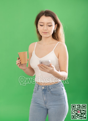 Woman standing on green background with smartphone and coffee in hands