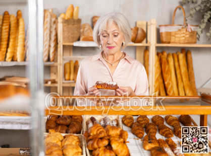 Senior female customer stands with croissant in hands near window of bakery.