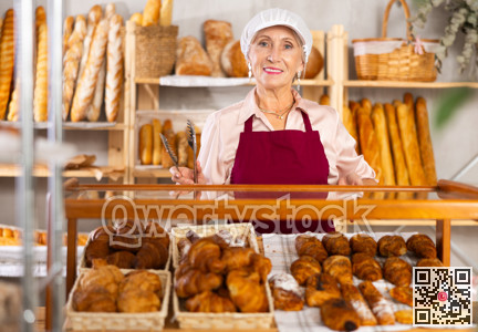 Portrait of elderly woman baker standing behind display case with fresh croissants. Offering to buy fresh baked goods