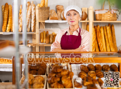 Portrait of elderly woman baker standing behind display case with fresh croissants. Offering to buy fresh baked goods