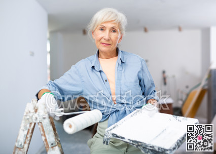 Mature woman painting walls with paint roller in refurbished room