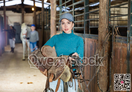 Woman jockey with saddle and bridle in her hands in stable