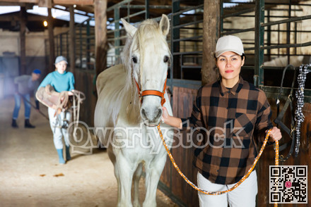 Asian woman leads a white horse by the bridle from stable