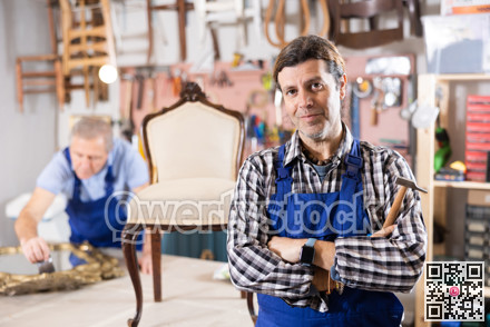 Portrait of middle-aged skillful repairman carpenter posing with crossed hands during workday in furniture woodwork studio