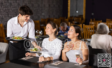 Young waiter serving ordered dishes to women in restaurant