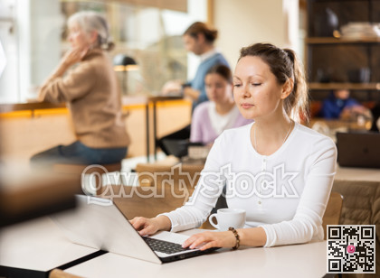 Positive European businesswoman drinking and enjoying coffee while working on laptop in cafe. Freelance and remote work