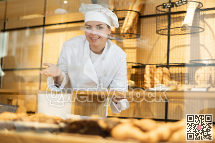 Female baker in a white coat and chef hat invites you to buy fresh bread and buns