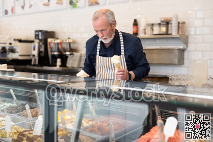 Elderly man in an apron offers ice cream at dessert counter with an inviting smile