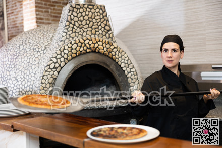Latino female chef in black uniform holding big shovel with pizza in kitchen