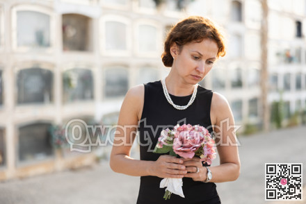 Sad woman in black dress with bouquet of flowers and handkerchief in hands, stands in cemetery