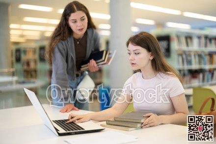 Two girls studying for an exam together in university library using books and laptop